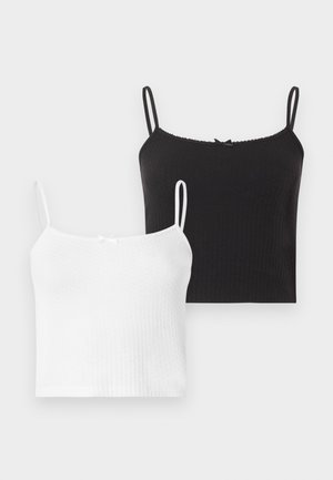 Jeune femme souriante, portant un top court noir, un jean noir à jambes larges et des chaussures noires à bouts pointus, debout devant un fond blanc.