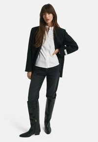 Blazer nero sopra una camicia bianca a bottoni, abbinato a jeans scuri e stivali neri lucidi fino al ginocchio con cerniere.