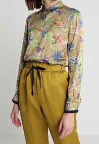 Blusa floreale in beige con motivi multicolori, con bottoni sul fronte e maniche lunghe. Abbinata a pantaloni giallo senape con vita con cordino.