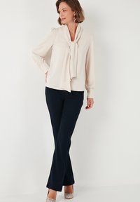 Beige blouse met een geknoopte strik bij de nek, gathered mouwmanchetten, gecombineerd met navy aansluitende broek en zilveren hoge hakken.