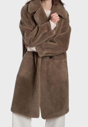 Femme portant un long manteau en fausse fourrure marron et pelucheux par-dessus un pull blanc, bras croisés, portant un pantalon blanc visible.