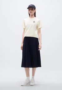 Ανοιχτό κίτρινο βαμβακερό μπλουζάκι με γεωμετρικό γραφικό, συνδυασμένο με πλισέ σκούρο μπλε φούστα και λευκά αθλητικά παπούτσια με μοναδικές υφασμάτινες λεπτομέρειες.