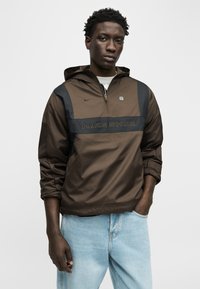 Νέος άνδρας με καφέ κοντό μπουφάν Nike Shox με κουκούλα και μαύρες λεπτομέρειες, φορώντας ανοιχτό μπλε τζιν, στέκεται μπροστά σε απλό λευκό φόντο.