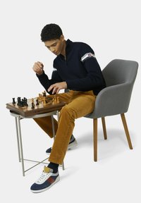 Marineblauw trui met ritssluiting en witte strepen, mosterdkleurige ribfluwelen broek, zittend in een grijze stoel, schaken aan een houten tafel. Sneakers met marineblauwe en rode accenten.