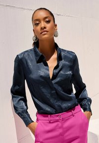 Marineblaues, glänzendes Langarmhemd mit feinem Muster, mit Knopfkragen und Tasche auf der Vorderseite, kombiniert mit leuchtend pinkfarbenen, taillierten Hosen.