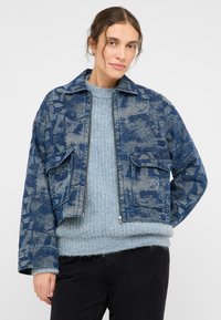 Veste en denim bleu à motifs avec col, fermeture éclair et deux poches avant. Portée sur un pull en laine bleu clair. La texture est visible.