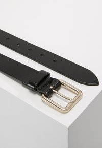Cintura in pelle nera con fibbia rettangolare in tono oro, diverse fori di regolazione e un'estremità affusolata. Texture liscia su tutta la superficie.