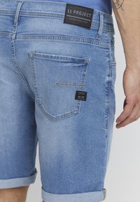 Lyse blå denimshorts med brett hem, med en baklomme og to merkedeapper som viser "11 PROJECT" og "20/20."
