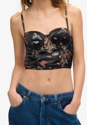 Femme portant un bustier court en satin noir avec des broderies colorées de dragon et de fleurs, associée à un jean en denim bleu.