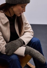 Moufles en laine texturée marron avec une forme allongée, portées avec une veste beige. Jeans foncés visibles, avec une simple chaise en bois en arrière-plan.
