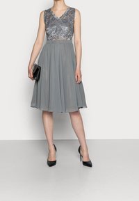 Robe grise avec un corsage en dentelle, un décolleté en V et une jupe fluide. Associée à des talons hauts noirs et une petite pochette noire.