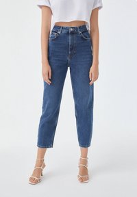 Jeans en denim bleu taille haute avec une coupe ample, présentant une fermeture à bouton, cinq poches et un ourlet retroussé. Portés avec un haut court blanc.