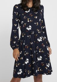 Robe florale marine à manches longues, taille cintrée et détail à nouer, présentant des motifs de fleurs blanches, jaunes et bordeaux sur un tissu doux.