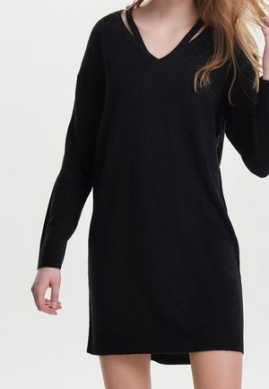 Vrouw draagt een zwarte, gebreide jurk met V-hals en lange mouwen, staand tegen een effen lichte achtergrond.