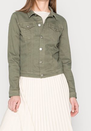 Vrouw draagt een olijfgroene spijkerjas met knopen over een gestreept shirt en een witte geplooide rok, houdt de rok met één hand vast.