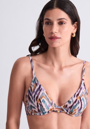 Femme portant un haut de bikini coloré à motifs avec des fines bretelles et des boucles d'oreilles en or, regardant légèrement sur le côté sur un fond clair.