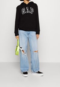 Sweat-shirt à capuche noir avec logo "GAP" blanc, associé à un jean large bleu clair délavé et usé. Tenant un petit sac à main vert, baskets noires.