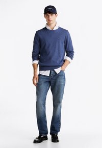 Mörkblå tröja, vit skjorta med krage under, blå jeans och svarta skor. Keps med logotyp, stående pose mot en enfärgad bakgrund.