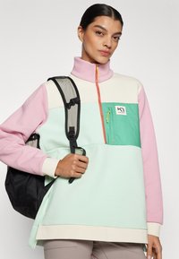 Farveblokeret pullover i pink, mint og creme, med høj krave, lynlås foran og en brystlomme med lynlås. Bærer en sort rygsæk.