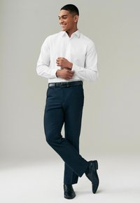 Weißes Langarmhemd und taillierte marineblaue Hose mit einem schwarzen Gürtel. Das Model steht in einer entspannten Pose vor einem hellgrauen Hintergrund. Schwarze Schuhe.