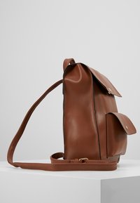 Ruskea nahkainen reppu, jossa on etutaskut ja säädettävät olkahihnat, esillä valkoisella alustalla yksinkertaisen taustan edessä.