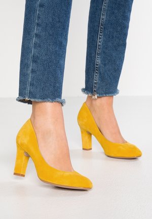 Gule suede sko med høje hæle, rundet tå og kraftig blokhæl. Teksturen er blød, og hælen måler cirka 7 cm.