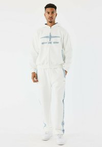 Witte hoodie en joggers met lichtblauwe denimaccenten. Voorzien van een ritssluiting aan de voorkant en een logo. Het materiaal lijkt zachter en casual.