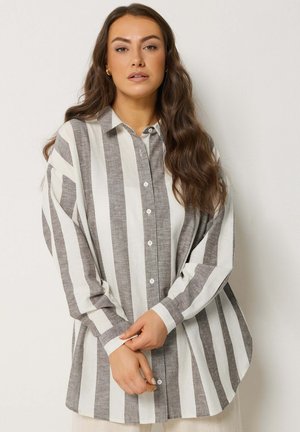 Vrouw met lang golvend haar die een oversized grijs-wit gestreept overhemd met verticale strepen draagt, met de handen gevouwen voor zich, tegen een effen achtergrond.