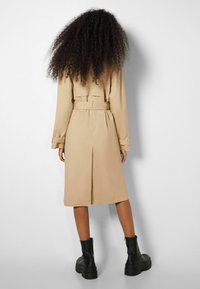 Manteau beige à ceinture à la taille, à manches longues et avec une fente au dos. Associé à des bottines noires jusqu'à la cheville avec une semelle épaisse.