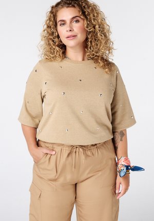 Femme aux cheveux bouclés portant un haut beige à manches courtes avec de petits clous et un pantalon beige à cordon assorti, avec une écharpe colorée attachée à son poignet.
