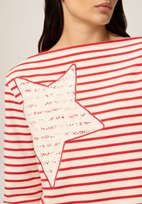 Mujer vestida con una blusa de rayas rojas y blancas con un gran aplique de encaje blanco en forma de estrella, contorneado en rojo, en el hombro y la espalda.