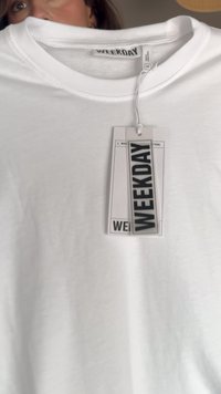 Bjela pamučna majica kratkih rukava s okruglim izrezom; ima pravokutnu oznaku s natpisom "WEEKDAY" ispisanim podebljanim, crnim slovima. Mekana tekstura i jednostavan dizajn.