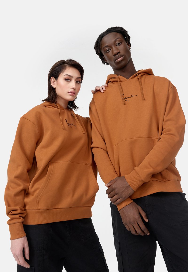 Sudaderas marrones con cordones y bolsillos frontales, con un logo en script negro. Modelos vistiendo pantalones negros, mostrando un ajuste relajado.