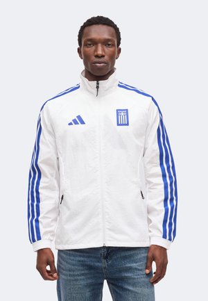 Uomo che indossa una giacca bianca con zip con strisce blu sulle maniche, logo Adidas ed emblema greco, abbinata a jeans blu.