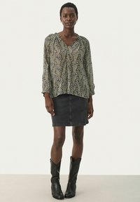 Svart blommönstrad blus med långa ärmar och V-ringning, som bärs med en svart denimkjol och höga svarta cowboystövlar. Enfärgad bakgrund.