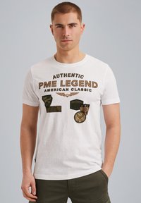 T-shirt en coton blanc avec des patches imprimés et du texte marron, arborant la marque "PME Legend" et des détails subtils sur le devant.