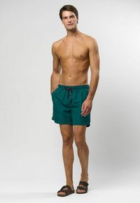 Vīriešu zaļie peldšorti, izgatavoti no viegla materiāla, ar elastīgu jostu, aukliņu un sānu kabatām. Valkāti ar brūnām čībām.