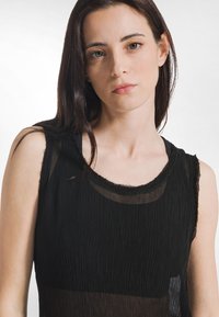 Czarny bezrękawnik o fakturowanym, plisowanym wzorze. Posiada dekolt w kształcie litery U oraz przezroczyste akcenty z tkaniny wzdłuż krawędzi.
