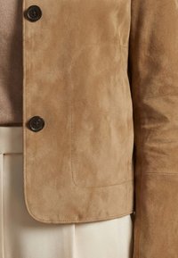Gros plan d'une veste en daim couleur sable avec des boutons foncés, portée sur un pantalon beige clair et un pull assorti beige.