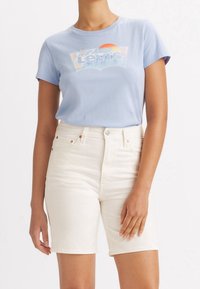 Kobieta ubrana w jasnoniebieską koszulkę Levi's z graficznym motywem zachodu słońca oraz białe szorty dżinsowe z wysokim stanem, stojąca na białym tle.