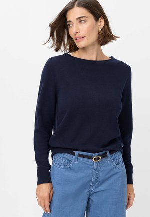 Kvinde med kort brunt hår iført guldringeøreringe, en mørkeblå sweater og lyseblå kordfløjlsbukser med sort bælte.