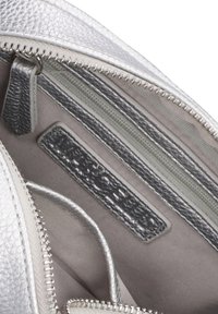 Interno di una borsa in pelle argentata con texture, dotato di più scomparti, inclusi sezioni con cerniera e un'etichetta con branding in rilievo.