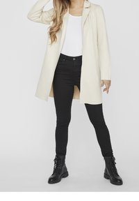 Kermanvärinen pitkä takki, jossa on lovikaulus, yhdistetty mustiin kapealahkeisiin farkkuihin ja mustiin nyörikenkiin. Takissa on suoraleikkainen malli.