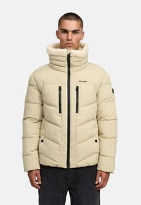 Bež zimska jakna s visokim ovratnikom, flis podstavom i dva crna džepa s patentnim zatvaračem na prednjoj strani. Ima chevron šavove i detalj s logotipom.