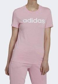 Růžové bavlněné tričko s krátkým rukávem a kulatým výstřihem, s velkým bílým logem "adidas" na přední straně. Hladká textura.