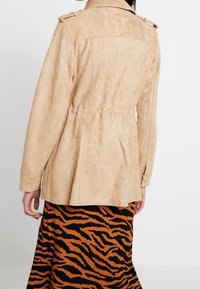 Giacca in suede beige con un design strutturato e tasche laterali, caratterizzata da passanti sulle spalle e vita stretta. Indossata sopra una gonna a fantasia.