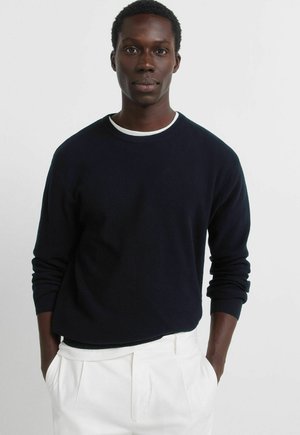 Granatowy prążkowany sweter dziany z długimi rękawami i zaokrąglonym dekoltem, noszony na białej koszuli. Gładka faktura i luźny krój.