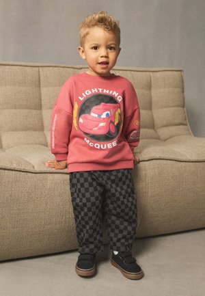 Lille barn står indendørs iført en rød Lightning McQueen sweatshirt og sorte ternede bukser, lænende sig op ad en beige sofa.