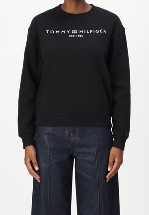 Zwarte sweater met lange mouwen met witte tekst "Tommy Hilfiger EST. 1985" over de borst, gecombineerd met donkerblauwe wijde jeans.