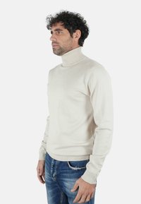 Uomo con capelli ricci che indossa un maglione a collo alto color crema e jeans blu, in piedi di profilo contro uno sfondo bianco uniforme.